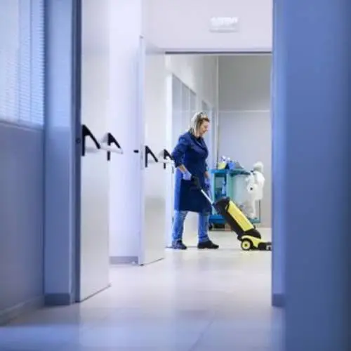 A woman in a blue coat and gloves cleans a hallway in a commercial or industrial cleaning setting.