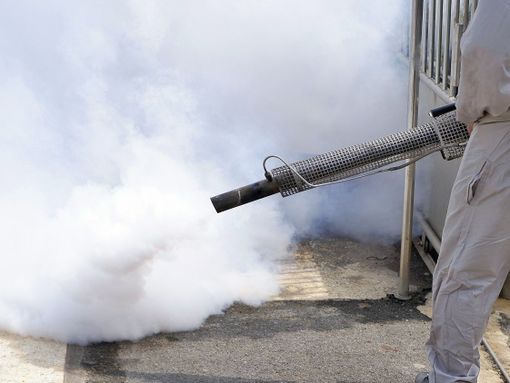 A person in a white coat releasing a dense cloud of smoke into the air using a spraying device.