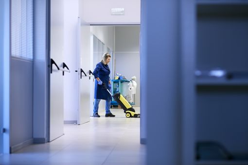 A woman in a blue coat diligently cleans a hallway, ensuring its cleanliness and tidiness.